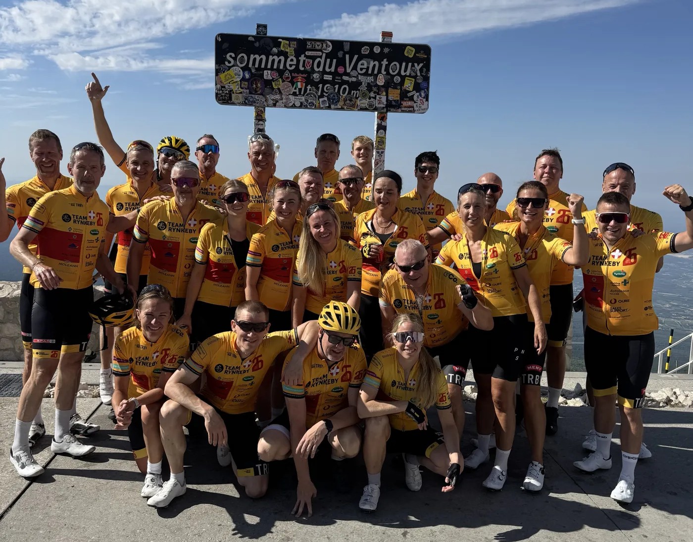 Interview Mark A team of cyclists in yellow jerseys celebrates at the summit of Mont Ventoux, posing in front of a sign under a blue sky.
