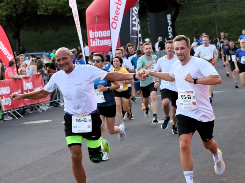 People running a race, two men in front holding hands, wearing white shirts with numbers, surrounded by other runners and spectators, red banners in background.