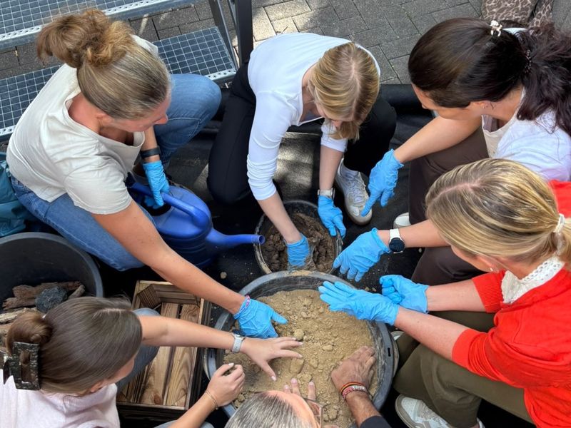 A group of people wearing blue gloves work together, mixing soil in a large container. One person is holding a blue watering can.
