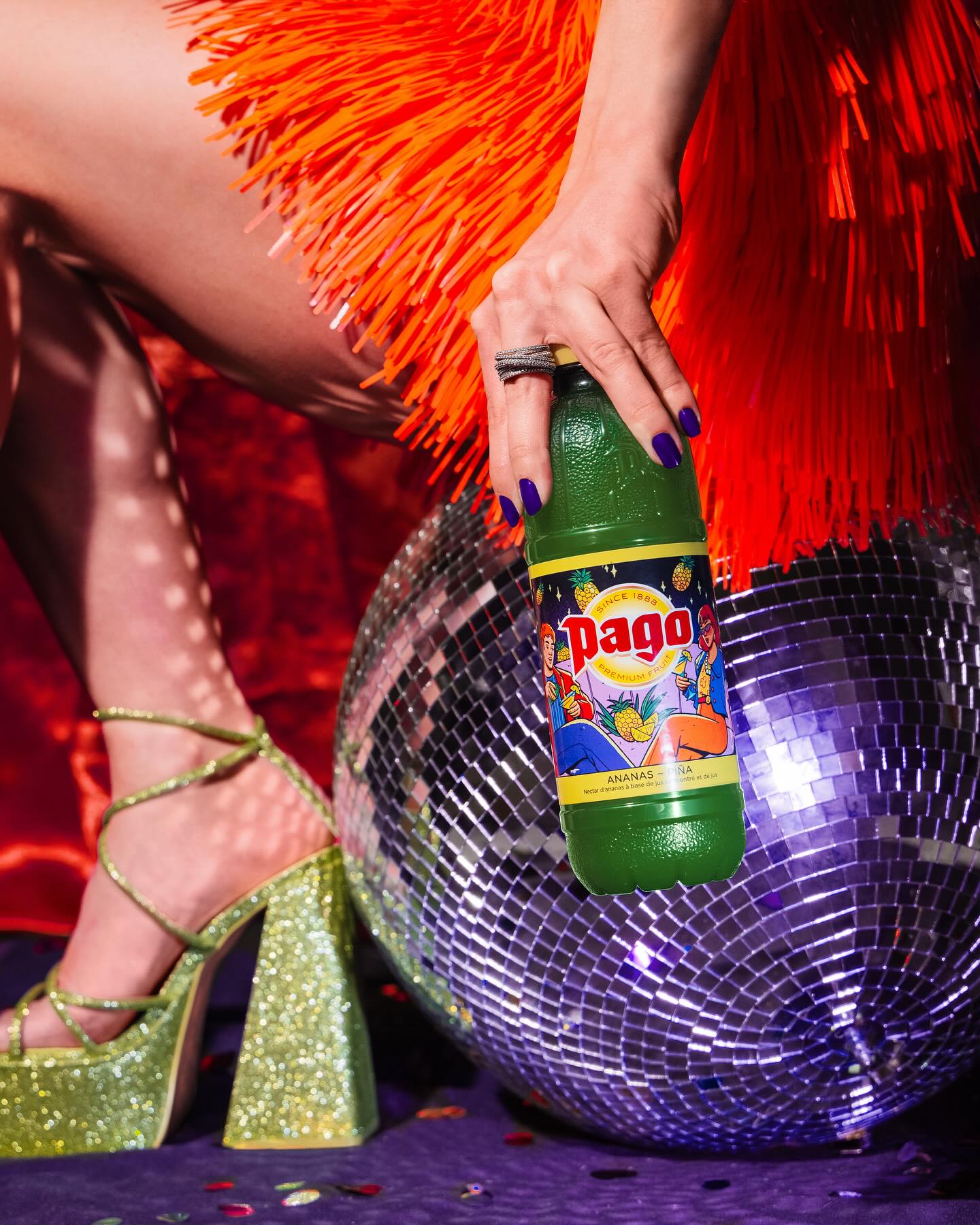 A person in an orange fringe outfit and glittery green heels holds a Pago bottle near a disco ball on a colorful background.