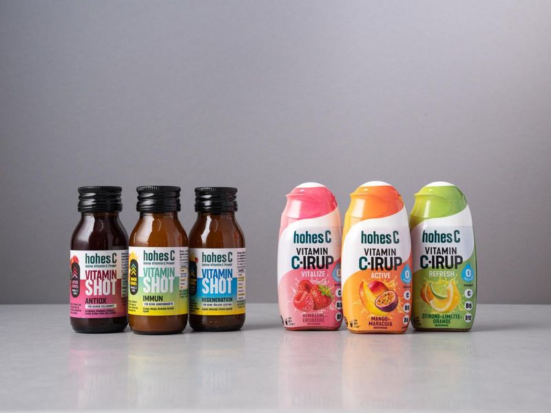 Six bottles of Hohes C vitamin drinks aligned on a surface, featuring Vitamin Shot and Vitamin C-irup varieties in various flavors.