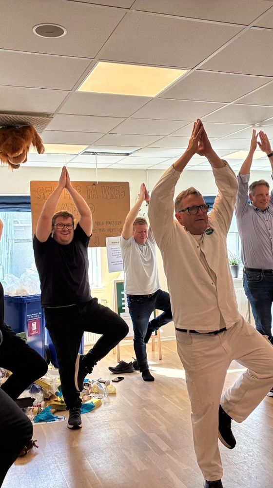 A group of people standing on one leg with arms raised in a yoga pose inside an office with a relaxed atmosphere.