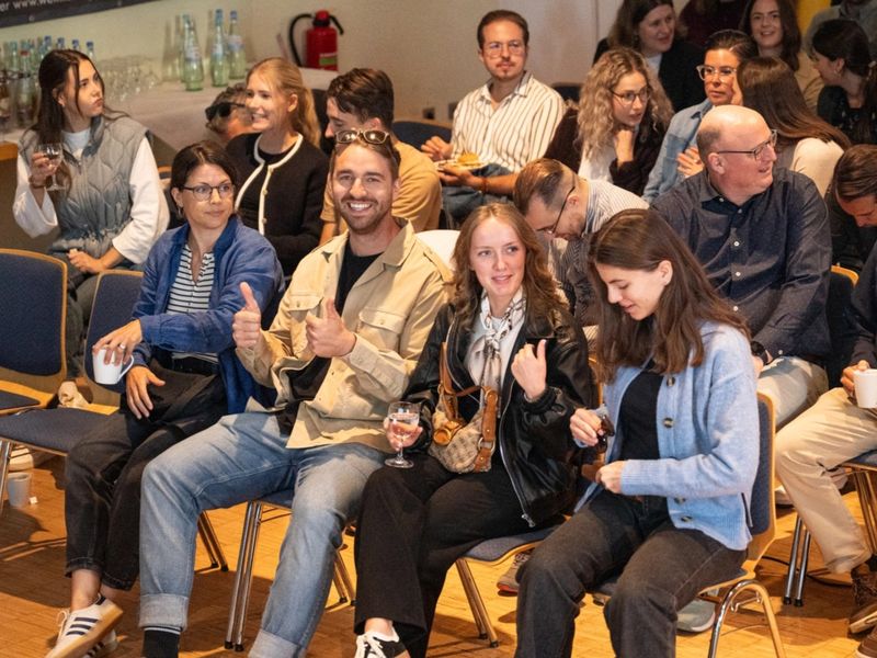 A group of people sitting in rows at an indoor event, with one person giving a thumbs-up and others engaging in conversation.