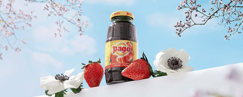 Bouteille de jus Pago Fraise posée entre des fraises fraîches et des fleurs sous un ciel bleu.