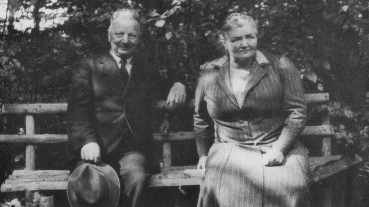 An elderly man and woman sit on a wooden bench in a garden, dressed in formal attire, with trees in the background.