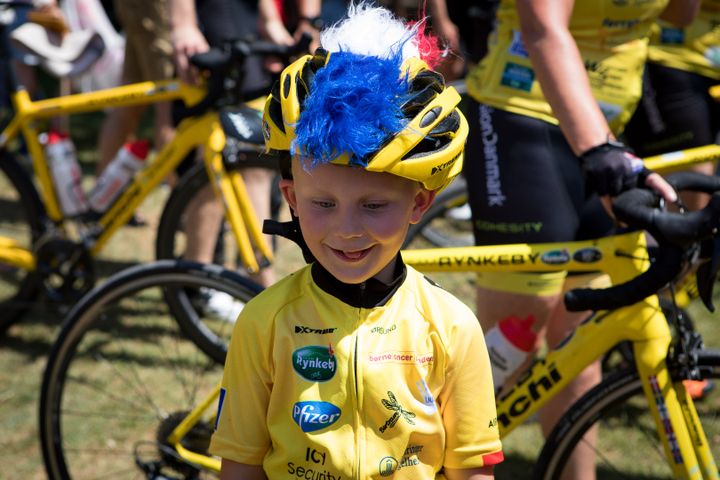 Child in a yellow cycling outfit and helmet with blue and red feathers, standing in front of bicycles.