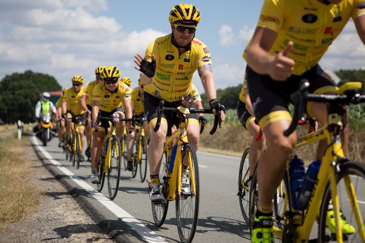Cyclists in yellow jerseys ride in formation on a rural road, with one rider waving and others giving a thumbs-up.