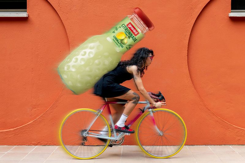 A woman rides a bike with a large bottle of granini Citronnade on her back, against an orange wall.