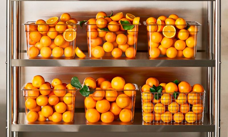 Plastic bins filled with fresh oranges and orange slices are neatly arranged on metal shelves. Some bins include green leaves for decoration.
