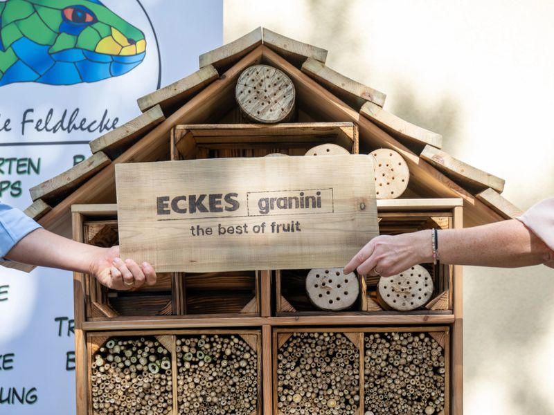 Two people hold a wooden sign with "ECKES granini, the best of fruit" in front of a wooden insect hotel, with a colorful poster in the background.