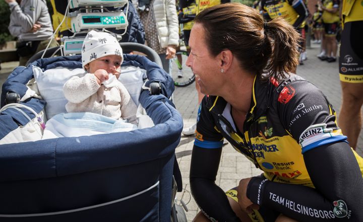 Cyclist in team attire kneels smiling at a baby lying in a stroller, surrounded by more cyclists.