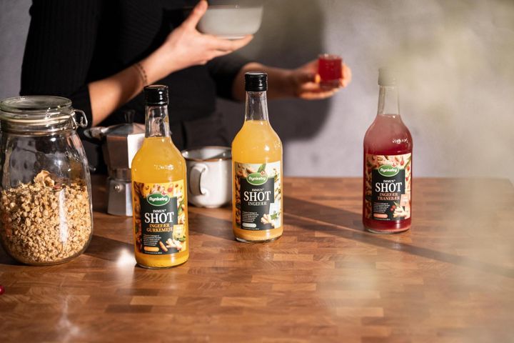 Bottles of ginger shots and granola jar on a wooden table. Person holds a bowl and a glass, blurred background.