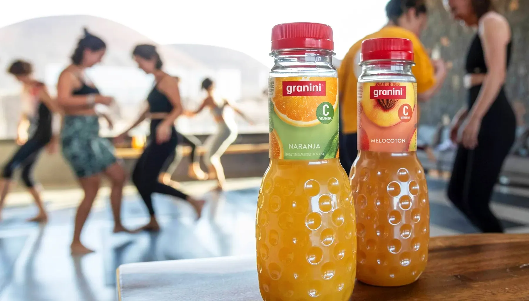 Dos botellas de zumo granini, una de naranja y otra de melocotón, sobre una mesa con un grupo de personas bailando al fondo, borroso.
