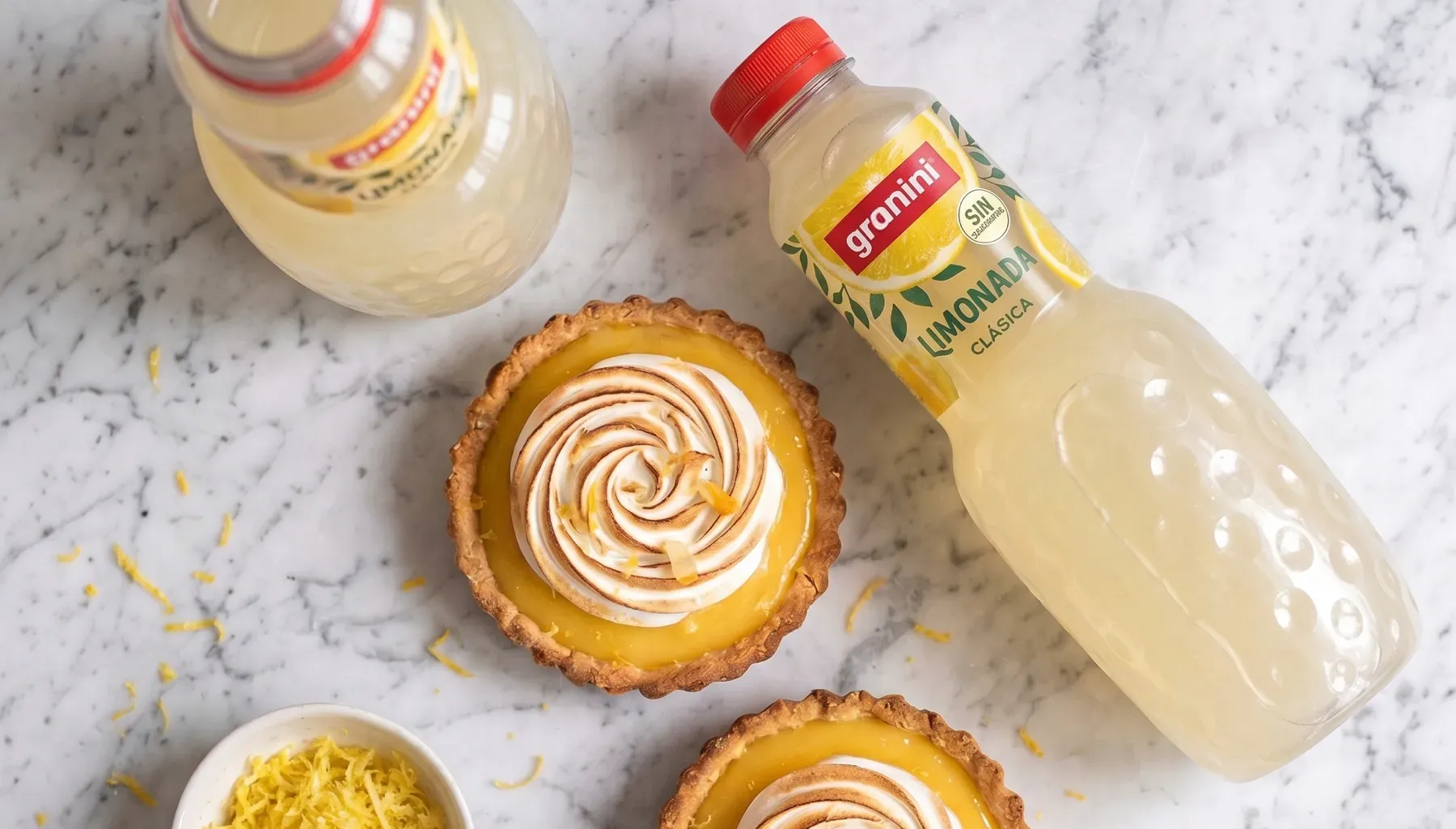 Dos tartaletas de merengue de limon sobre una superficie de marmol, rodeadas de ralladura de limon y botellas de granini Limonada Clasica.