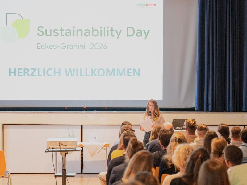 A woman stands at a podium speaking to an audience. A large screen displays "Sustainability Day" and "Herzlich Willkommen" in German.