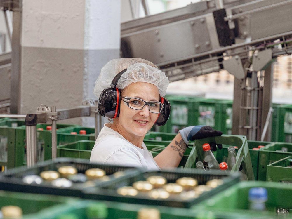 A factory worker wearing a hairnet, safety glasses, and ear protection smiles while handling crates of bottles in an industrial setting.
