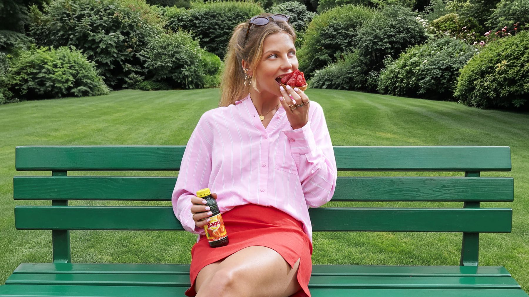 Femme assise sur un banc dans un jardin, dégustant une tarte aux fraises avec une bouteille de jus Pago.