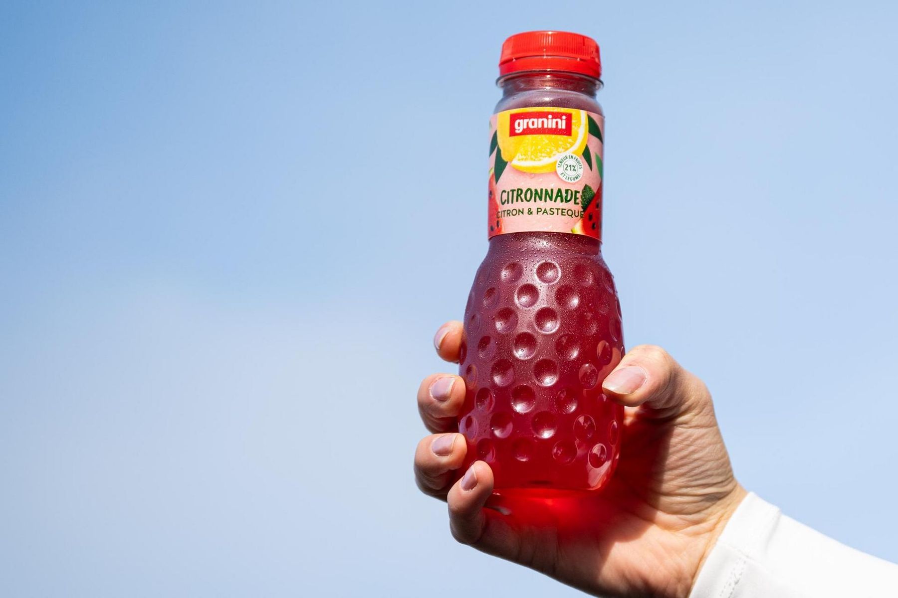 Hand holding a Granini citronnade bottle with red liquid against a clear blue sky.