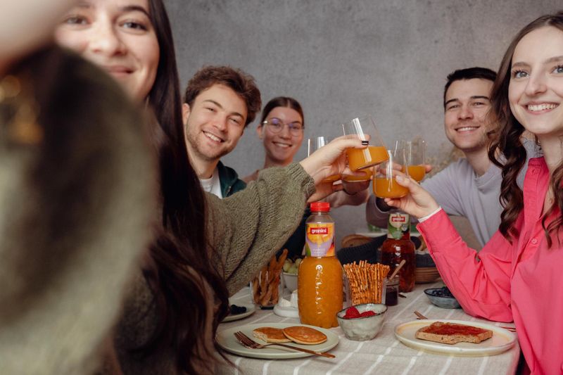 Group of friends cheers with orange juice at a table with breakfast items like pancakes and strawberries.