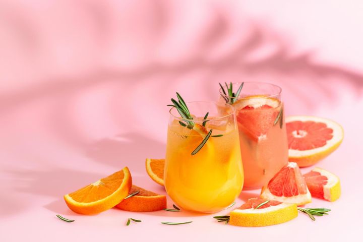 Two glasses of citrus cocktails with rosemary garnish, surrounded by sliced oranges and grapefruits, on a pink background with shadows.