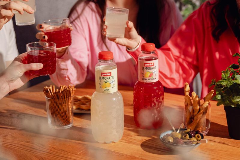 People holding glasses of lemonade and cranberry juice at a table with snacks, including pretzel sticks, breadsticks, and olives.
