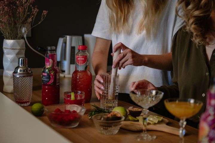Two people preparing cocktails at a kitchen counter with various ingredients, including fruit, bottles, and a cocktail shaker.