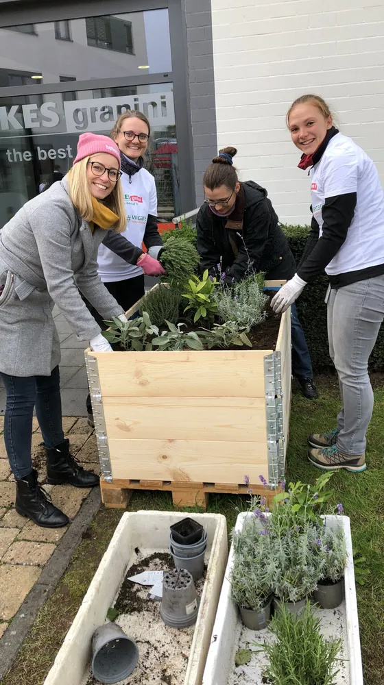 Four people are gathered around a wooden planter box, preparing to plant herbs and flowers. They are smiling, wearing casual clothing and gloves.