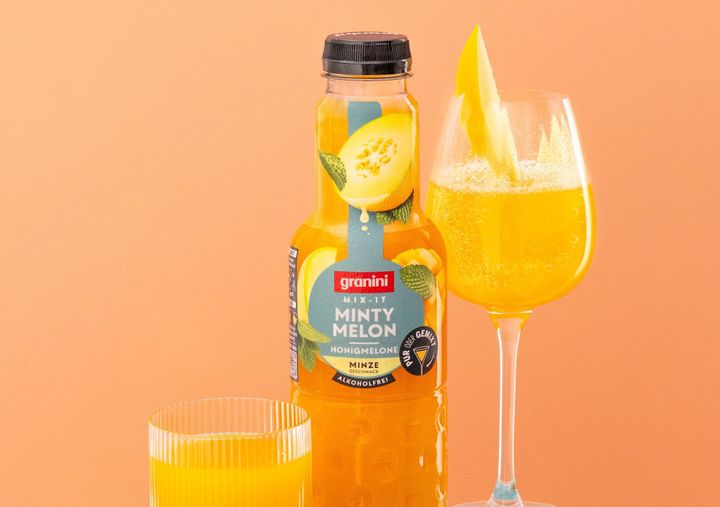 A bottle of Granini Minty Melon juice is next to a glass and a wine glass with orange juice on an orange background.