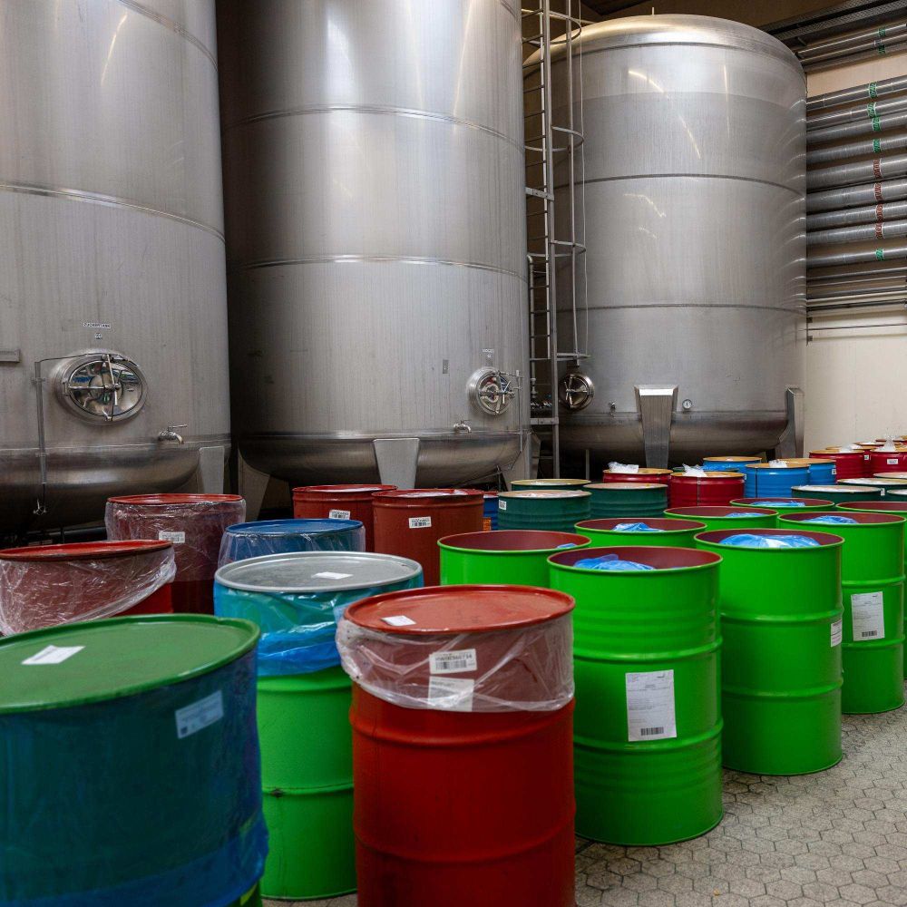 Colorful barrels are arranged in front of large stainless steel tanks inside an industrial facility, creating a vibrant contrast.