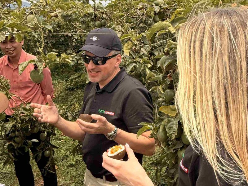 A man in sunglasses holding a halved passion fruit explains something to a group in a lush orchard, with green passion fruit visible in the background.