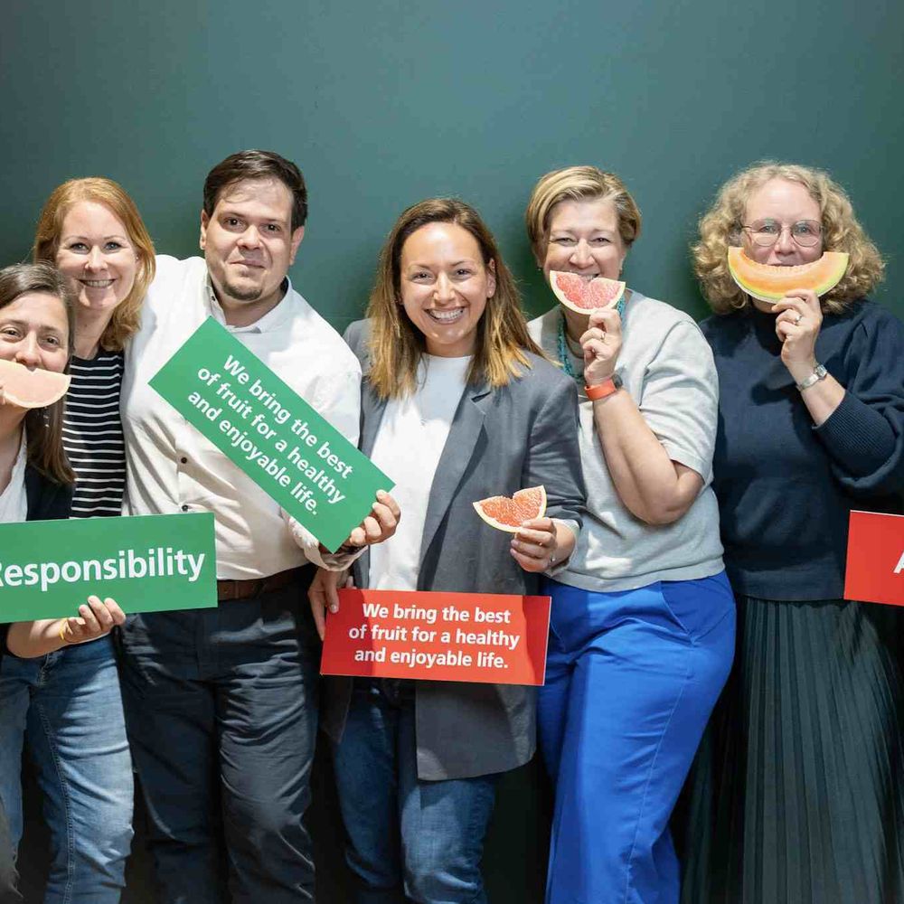 A group of seven people smiling, holding fruit slices and signs saying "Responsibility," "Ambition," and a message about healthy living with fruit.