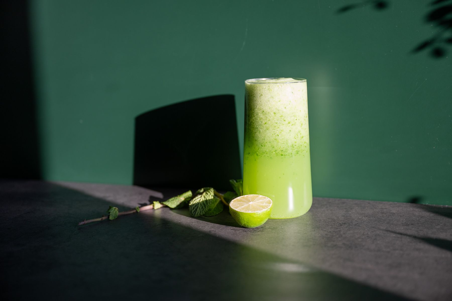 A tall glass of a Passion Fizz Cocktail with a frothy top, accompanied by a halved lime and mint leaves, set against a dark green background.