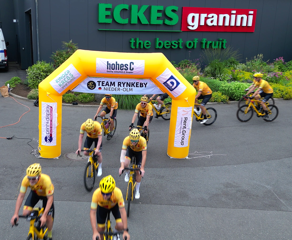 Cyclists in yellow jerseys ride under a yellow arch labeled "Team Rynkeby Nieder-Olm," with a building in the background displaying "Eckes Granini."