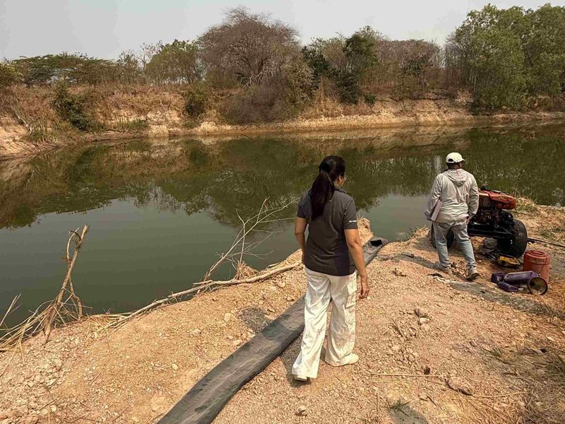 Two people are working near a pond surrounded by dry vegetation, with machinery beside them on a dirt embankment.