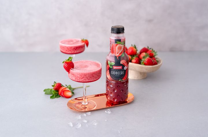Bottle of granini Erdbeere next to two glasses of pink drinks, fresh strawberries, and ice on a copper tray.