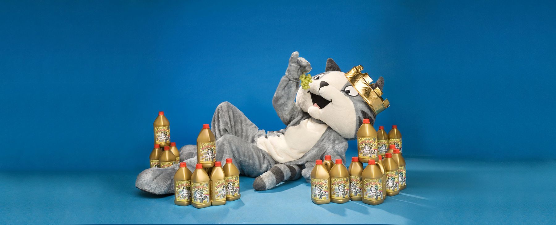 A costumed character in a crown lounges on the floor, surrounded by numerous gold bottles, against a bright blue background.