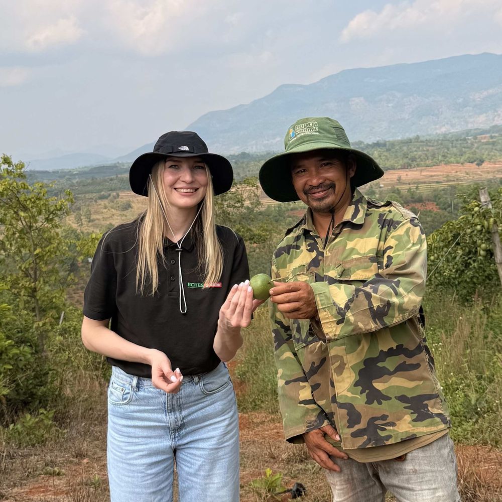Two people in sun hats smiling, holding a lemon in a rural landscape with mountains and greenery in the background.