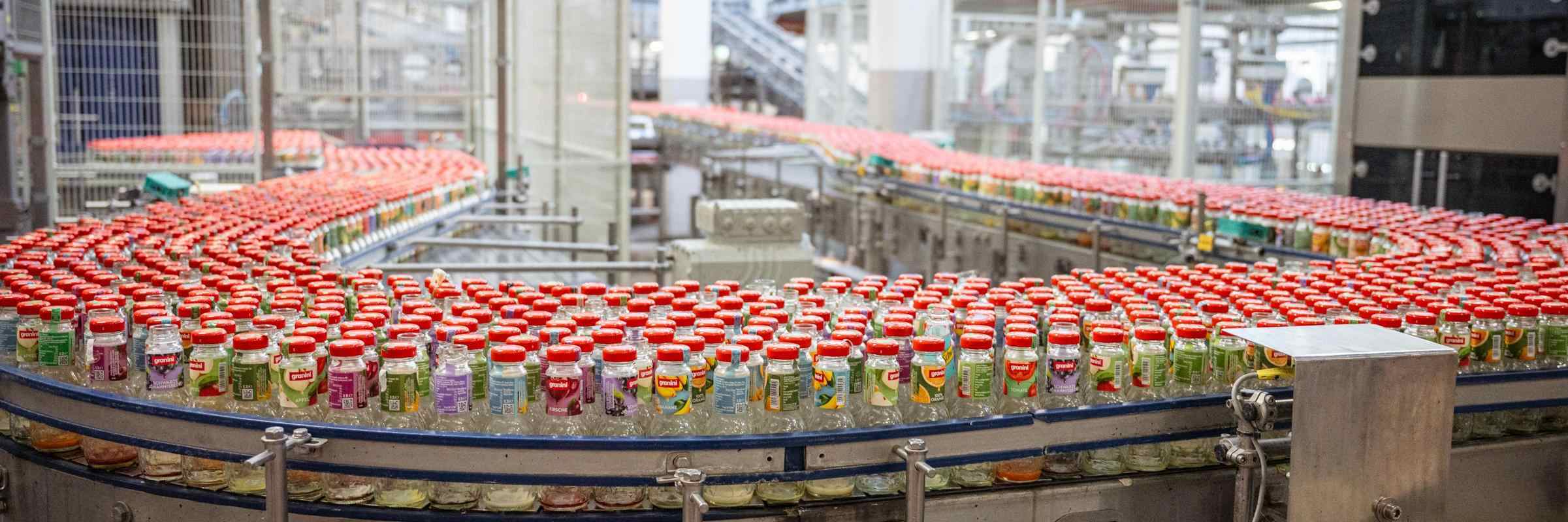A conveyor belt filled with rows of juice bottles with red caps in a factory setting.