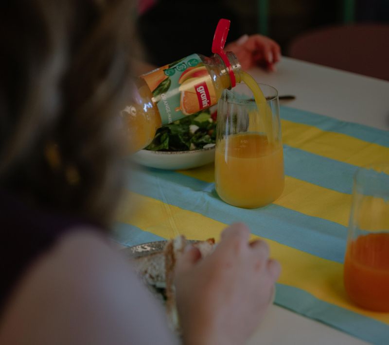 Person pouring orange juice into a glass on a table with a salad bowl and a colorful striped tablecloth.