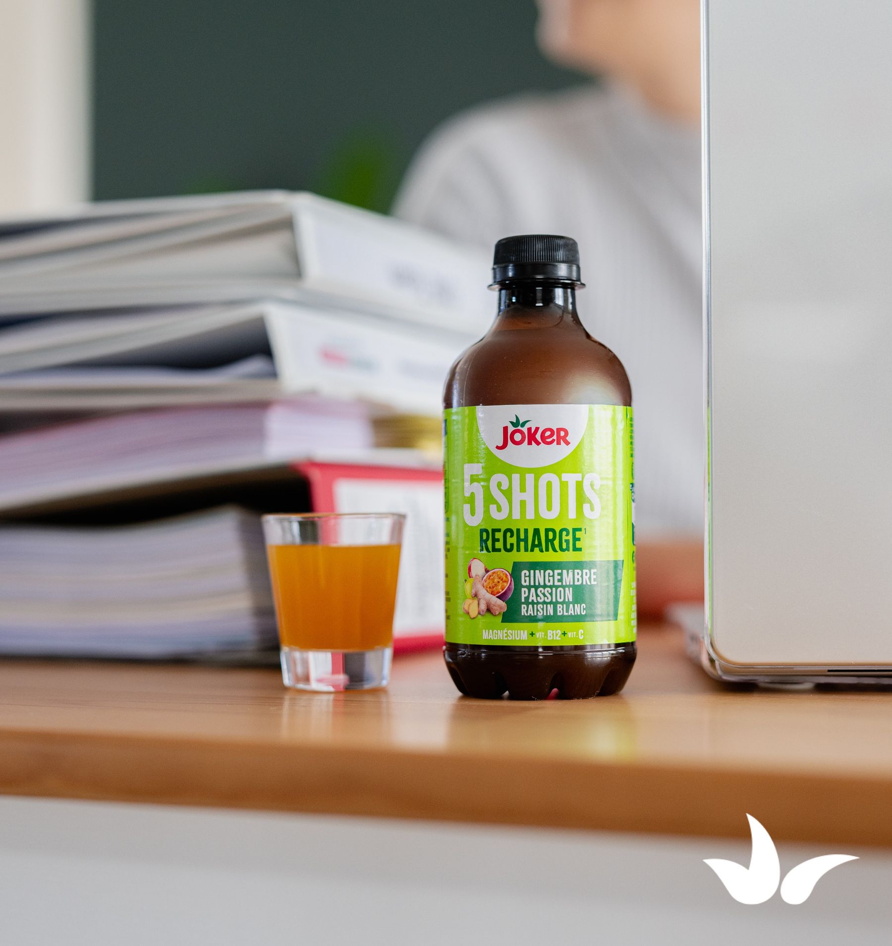 A bottle of Joker 5 Shots Recharge with ginger, passion fruit, and white grape flavor next to a glass of orange liquid on a wooden desk.