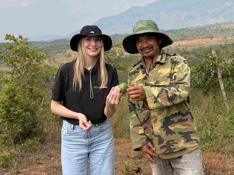 Two people in hats are holding a green fruit in a rural landscape with mountains in the background.