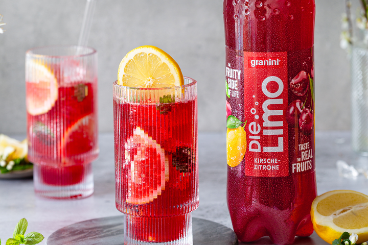 A glass of red fruit punch with lemon and mint, next to a bottle of Granini Die Limo, on a gray surface.