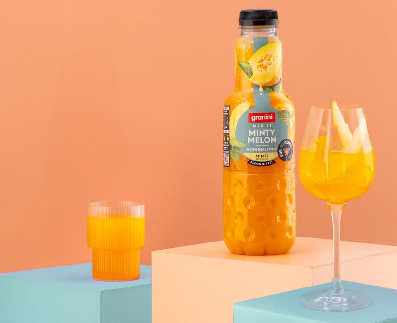 Minty Melon juice bottle on a pedestal, a filled glass with melon slices, and another glass against a peach background.