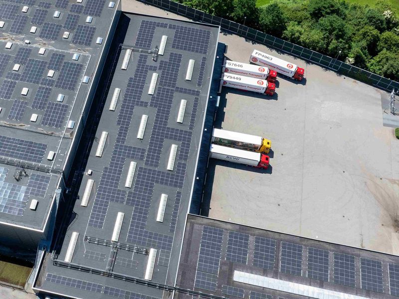 Aerial view of a large warehouse with solar panels on the roof and several red trucks parked in a loading bay area.