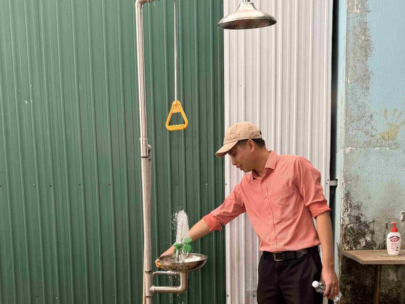 Man in a pink shirt and beige cap uses an emergency eyewash station, while holding a water bottle, in front of corrugated metal walls.