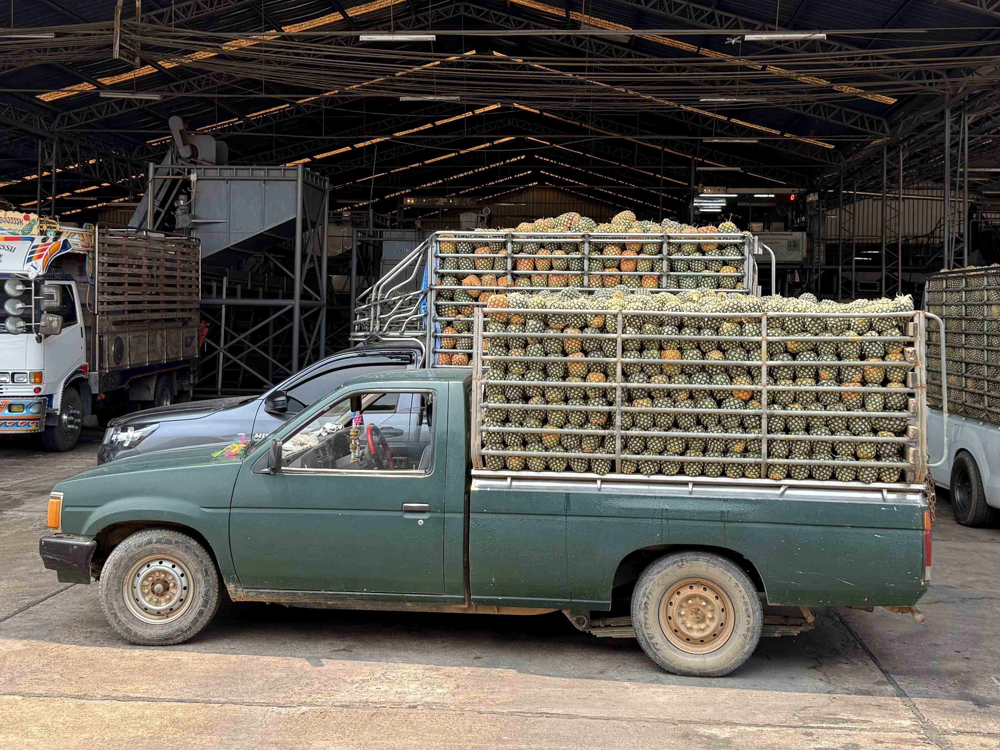 A green pickup truck loaded with pineapples is parked inside an industrial warehouse. Other vehicles and machinery are visible in the background.