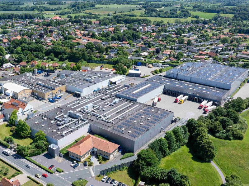Aerial view of a large industrial complex with multiple warehouse buildings, surrounded by green trees and a residential area in the background.