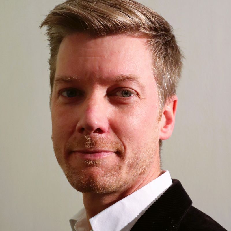 Man with short light brown hair and a trimmed beard, wearing a white shirt and black jacket, looking slightly to the side.