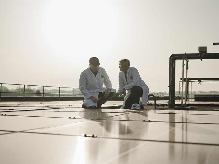 Two people in lab coats kneel on a rooftop, inspecting solar panels under a bright sunlit sky.