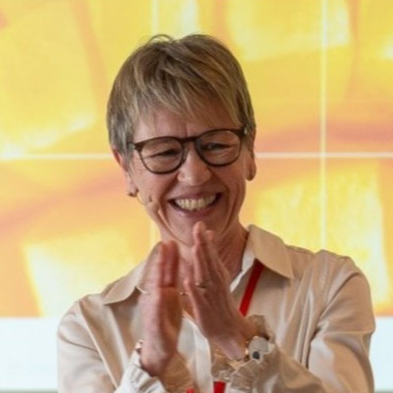 A smiling person with short hair and glasses clapping, wearing a white shirt and standing in front of a bright, abstract background.